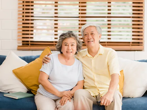 An older Asian couple sits close together on a sofa, smiling warmly as the man embraces his wife in a bright, cozy living room.
