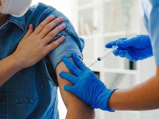 Masked patient in a denim shirt receives an upper-arm vaccine shot from a gloved clinician in a bright clinic, conveying a calm, reassuring medical moment.