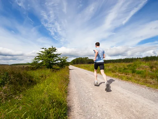 A lone runner in a blue shirt jogs along a sunlit gravel path through green countryside beneath a vast, streaky blue sky, evoking freedom and calm.