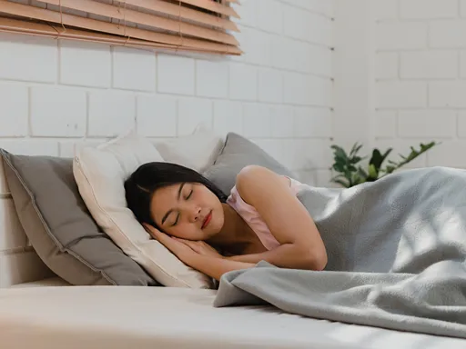 A woman sleeps peacefully on her side in a bright bedroom, nestled among white pillows under a soft gray blanket; daylight filters through wooden blinds.